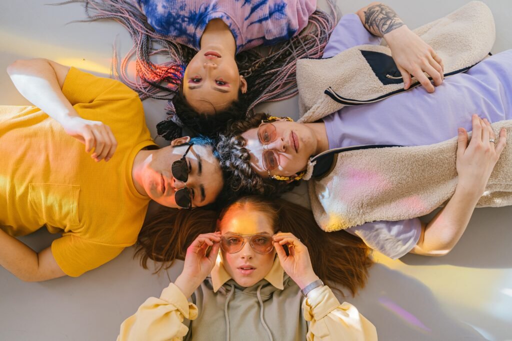 Diverse group of young friends posing on floor in colorful casual outfits and sunglasses.