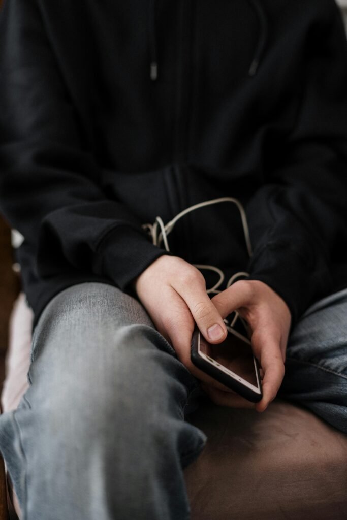 pexels-photo-4100483-4100483 Close-up of a teenager holding a smartphone, wearing a black hoodie, seated indoors.